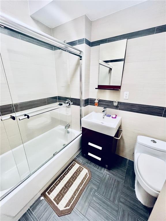 bathroom featuring shower / bath combination with glass door, tile walls, toilet, a sink, and tile patterned floors