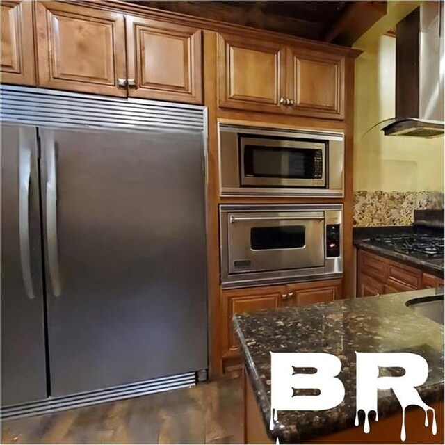 kitchen featuring wall chimney range hood, built in appliances, and dark stone countertops