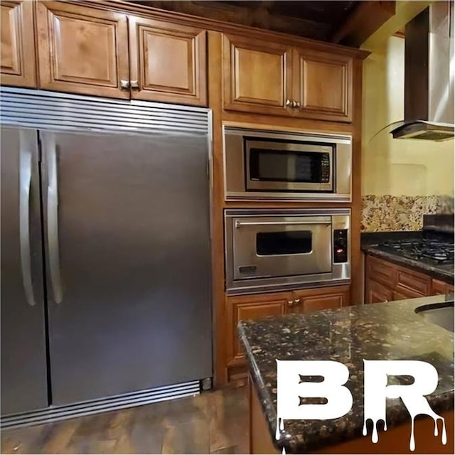 kitchen with wall chimney range hood, built in appliances, and dark stone countertops