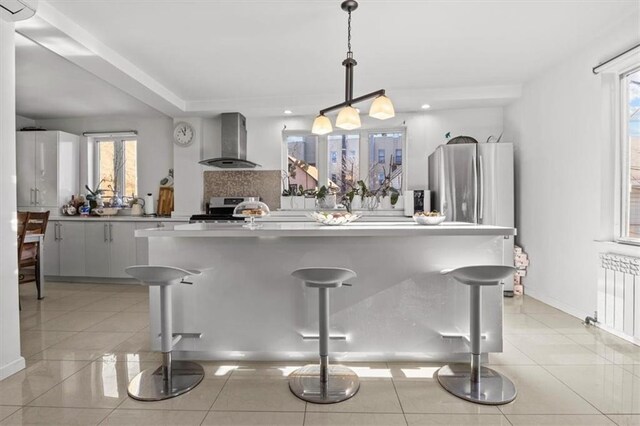 kitchen featuring a breakfast bar, hanging light fixtures, radiator, stainless steel appliances, and wall chimney exhaust hood