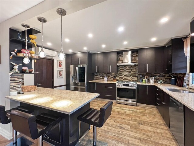 kitchen with light stone counters, appliances with stainless steel finishes, a kitchen breakfast bar, pendant lighting, and wall chimney range hood