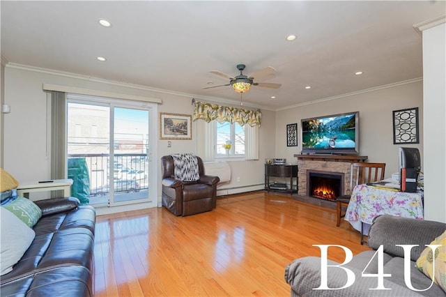 living room with ceiling fan, baseboard heating, hardwood / wood-style floors, ornamental molding, and a stone fireplace