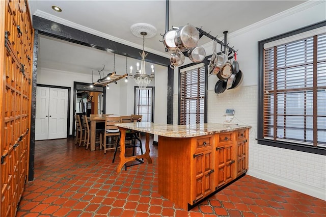 kitchen with a wainscoted wall, ornamental molding, light stone counters, brown cabinets, and hanging light fixtures