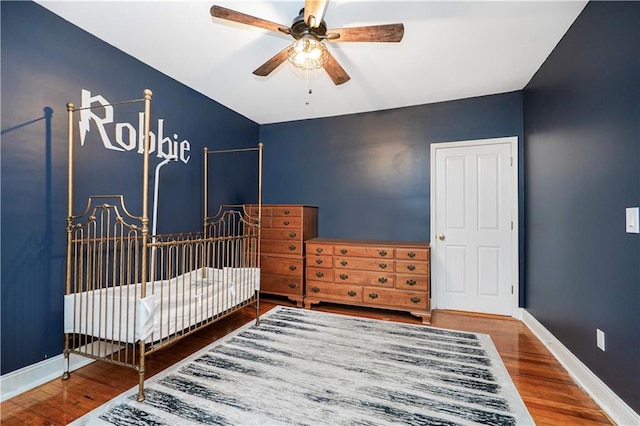 bedroom with a nursery area, ceiling fan, baseboards, and wood finished floors