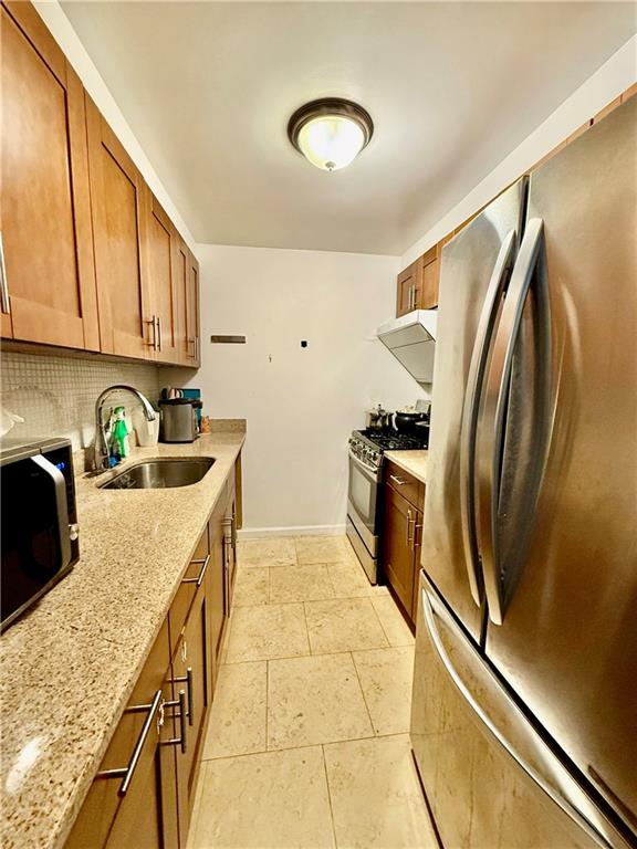 kitchen with a sink, tasteful backsplash, brown cabinets, light stone countertops, and appliances with stainless steel finishes