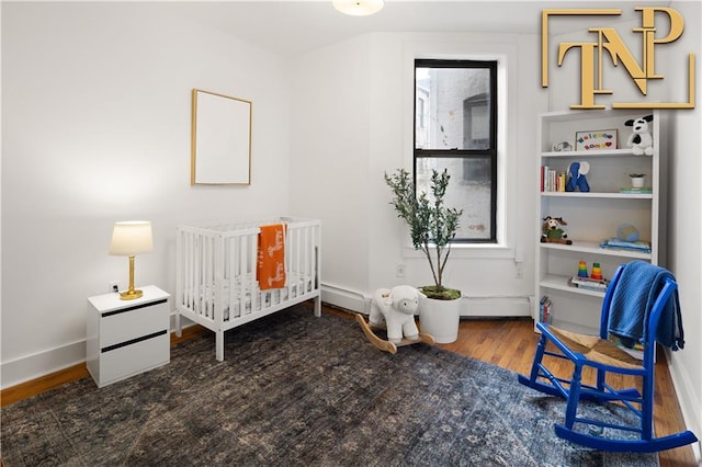 bedroom with a crib, a baseboard heating unit, baseboards, and wood finished floors