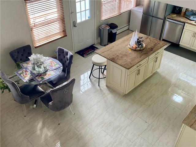 kitchen featuring appliances with stainless steel finishes, baseboard heating, cream cabinetry, light stone countertops, and washer / dryer