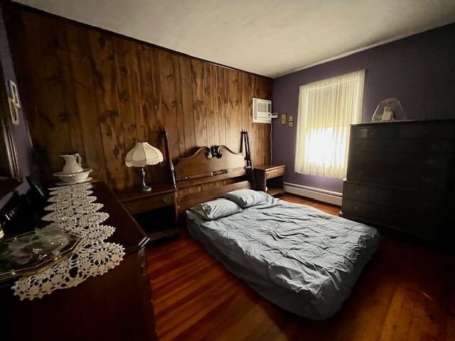 bedroom with dark wood-type flooring, a baseboard radiator, a wall mounted air conditioner, and wood walls