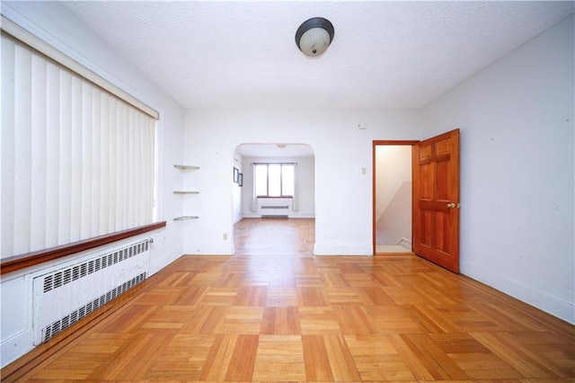 empty room featuring arched walkways, a textured ceiling, baseboards, and radiator