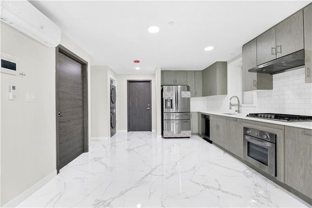 kitchen with stacked washer and dryer, gray cabinets, appliances with stainless steel finishes, a sink, and under cabinet range hood