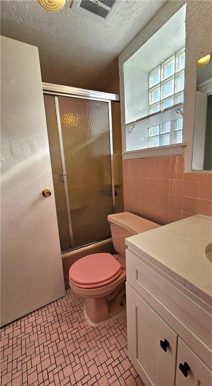 full bathroom featuring combined bath / shower with glass door, tile walls, vanity, a textured ceiling, and toilet