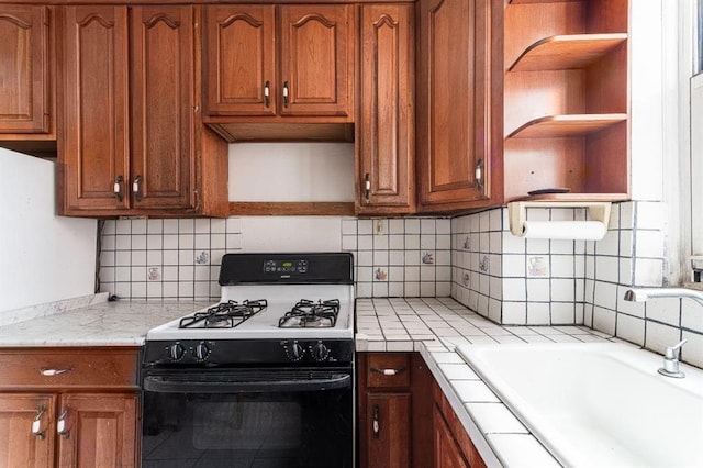 kitchen featuring brown cabinetry, open shelves, tile counters, gas range oven, and a sink