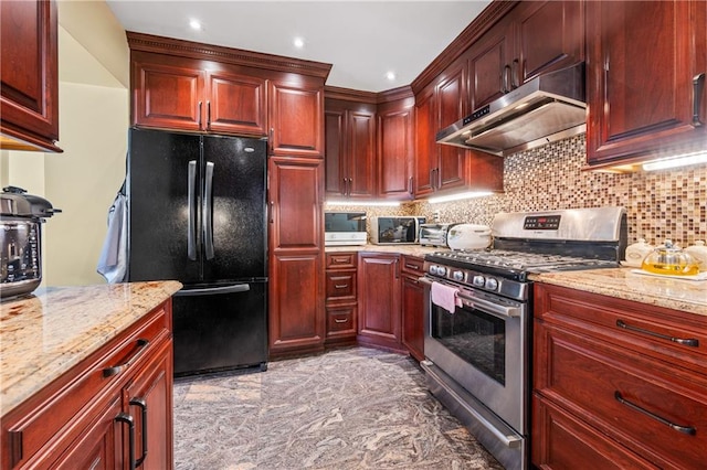 kitchen featuring under cabinet range hood, light stone counters, freestanding refrigerator, stainless steel range with gas cooktop, and dark brown cabinets