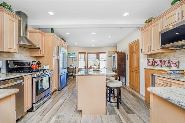 kitchen featuring stainless steel appliances, light brown cabinets, a kitchen island, wall chimney range hood, and light wood-type flooring