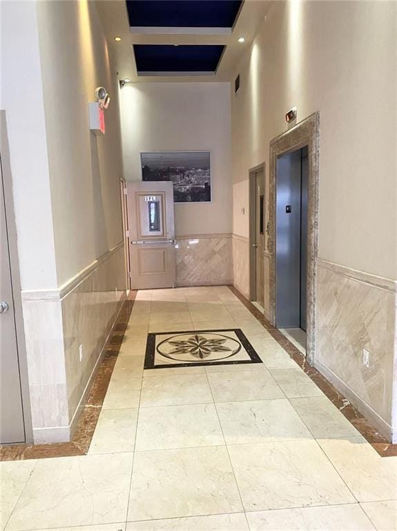 hall with a wainscoted wall, visible vents, a towering ceiling, tile patterned floors, and elevator