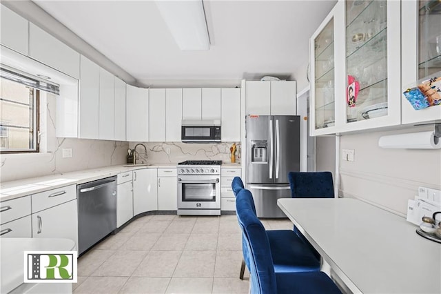 kitchen featuring stainless steel appliances, tasteful backsplash, glass insert cabinets, light tile patterned flooring, and white cabinets