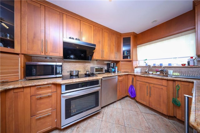 kitchen featuring light stone counters, a sink, appliances with stainless steel finishes, brown cabinets, and glass insert cabinets