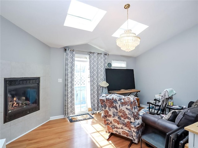 living area featuring a notable chandelier, a tiled fireplace, lofted ceiling with skylight, wood finished floors, and baseboards