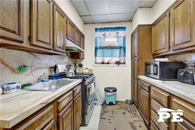 kitchen featuring a drop ceiling, under cabinet range hood, stainless steel appliances, a sink, and brown cabinets