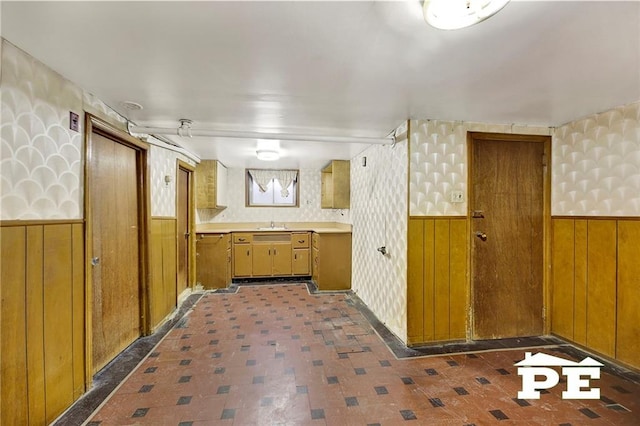 kitchen featuring brown cabinetry, light countertops, a wainscoted wall, and wallpapered walls