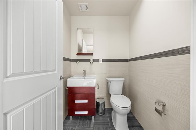 half bathroom featuring toilet, vanity, visible vents, tile walls, and wainscoting