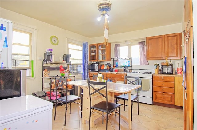kitchen with brown cabinets, light tile patterned flooring, stainless steel microwave, white range with gas cooktop, and glass insert cabinets