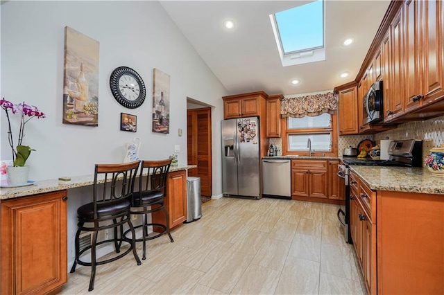 kitchen with light stone counters, stainless steel appliances, brown cabinetry, a sink, and vaulted ceiling with skylight