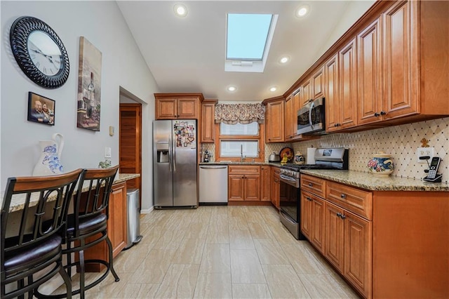 kitchen with a skylight, brown cabinetry, decorative backsplash, appliances with stainless steel finishes, and light stone countertops