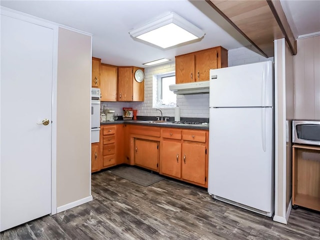 kitchen with white appliances, dark countertops, dark wood-style flooring, under cabinet range hood, and a sink