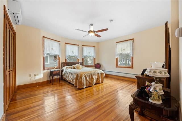 bedroom with a wall unit AC, light wood-style flooring, baseboard heating, ceiling fan, and baseboards