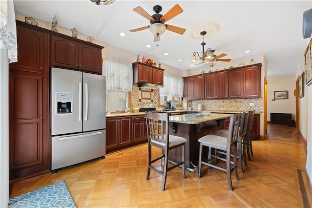 kitchen featuring light stone counters, a kitchen island, stainless steel fridge, under cabinet range hood, and a kitchen breakfast bar