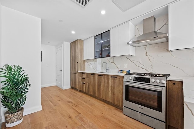 kitchen with stainless steel range with gas stovetop, wall chimney range hood, decorative backsplash, light wood-style flooring, and white cabinetry