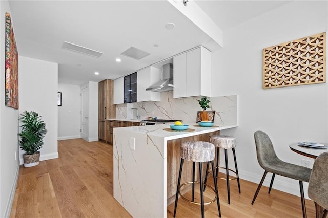 kitchen featuring light wood finished floors, a kitchen breakfast bar, wall chimney exhaust hood, a peninsula, and tasteful backsplash