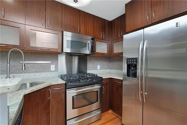 kitchen with light stone counters, stainless steel appliances, backsplash, glass insert cabinets, and a sink