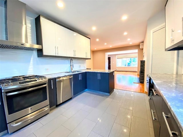 kitchen with wall chimney range hood, appliances with stainless steel finishes, a sink, a peninsula, and white cabinets