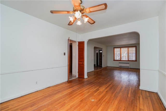 empty room featuring arched walkways, radiator, a ceiling fan, light wood-type flooring, and baseboards