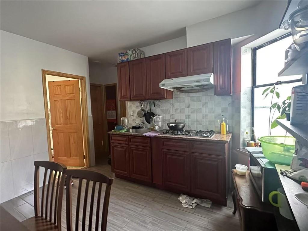 kitchen with a sink, light countertops, under cabinet range hood, stainless steel gas stovetop, and tile walls