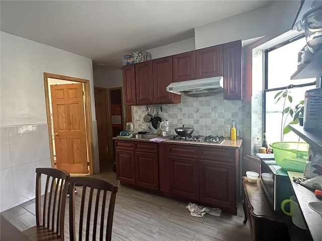 kitchen with a sink, light countertops, under cabinet range hood, stainless steel gas stovetop, and tile walls