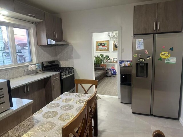 kitchen featuring under cabinet range hood, backsplash, stainless steel appliances, light stone countertops, and dark brown cabinets