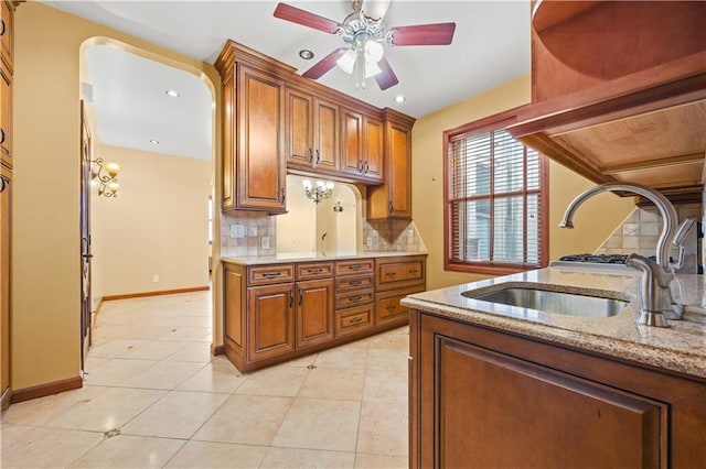 kitchen featuring brown cabinets, a ceiling fan, a sink, decorative backsplash, and light stone countertops