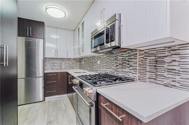 kitchen with stainless steel appliances, a sink, dark brown cabinets, decorative backsplash, and glass insert cabinets