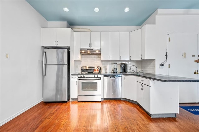 kitchen with dark countertops, under cabinet range hood, appliances with stainless steel finishes, a peninsula, and a sink