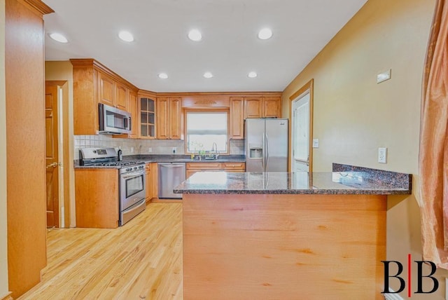 kitchen with light wood finished floors, glass insert cabinets, dark stone counters, a peninsula, and stainless steel appliances