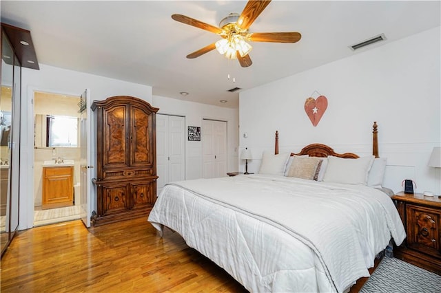 bedroom featuring a ceiling fan, visible vents, light wood finished floors, ensuite bath, and two closets