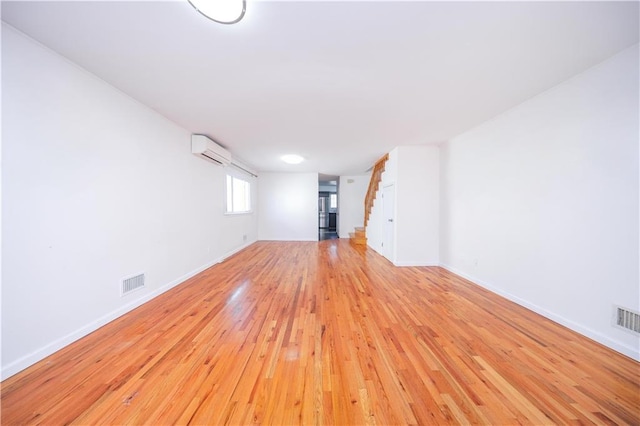 unfurnished living room with a wall unit AC, stairway, visible vents, baseboards, and light wood-style floors