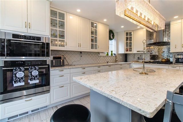 kitchen with light stone counters, double oven, a breakfast bar area, wall chimney exhaust hood, and decorative backsplash