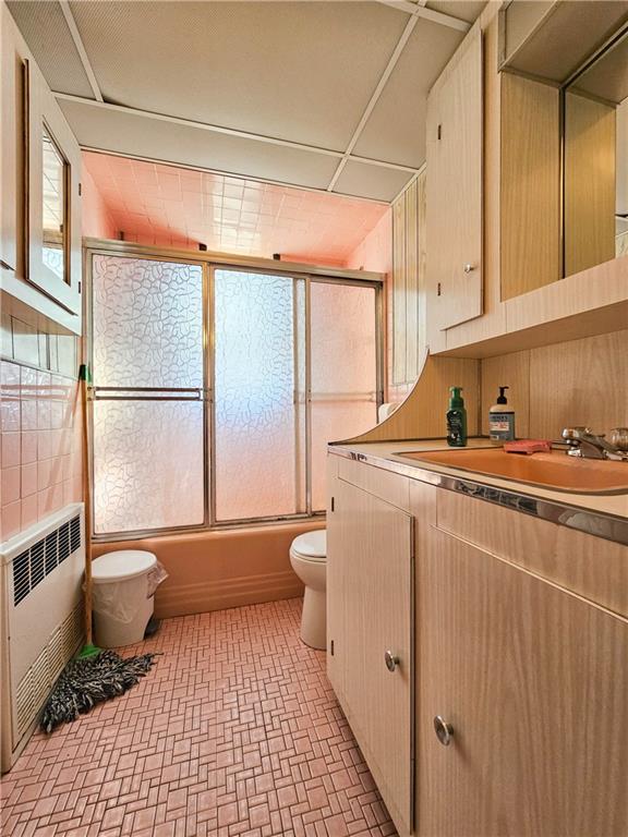 full bath featuring shower / bath combination with glass door, vanity, toilet, brick patterned floor, and radiator heating unit