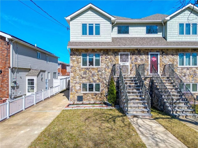 multi unit property with a shingled roof, fence, stairs, a front yard, and stone siding