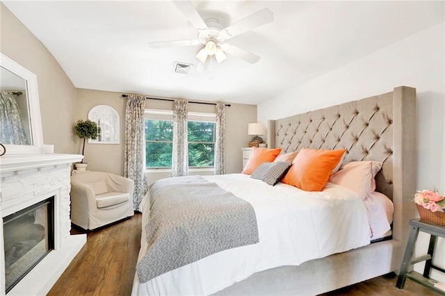 bedroom with a ceiling fan, visible vents, a stone fireplace, and wood finished floors