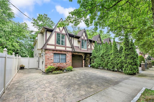 english style home featuring stucco siding, decorative driveway, fence, a garage, and brick siding